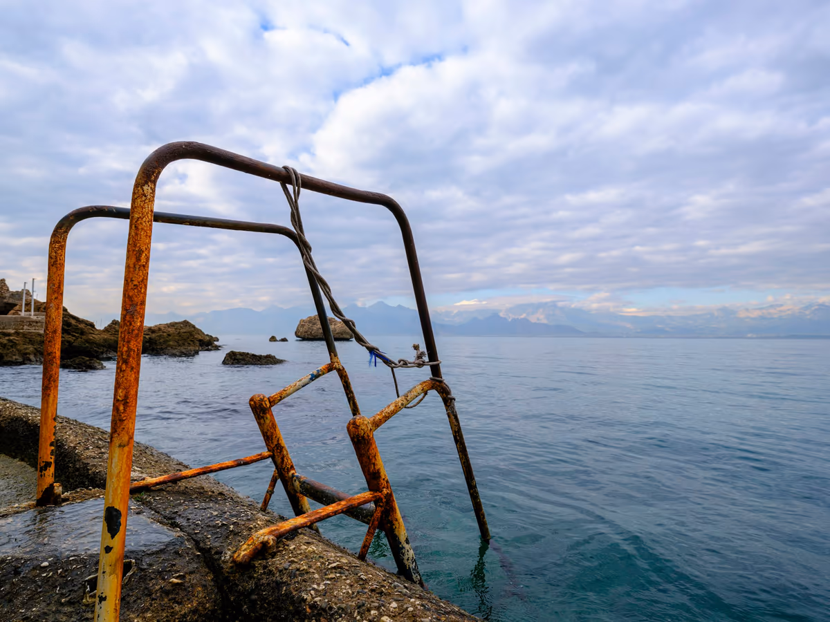 Garde-corps métallique fortement corrodé au bord de l'eau, tubes rouillés et scellements attaqués