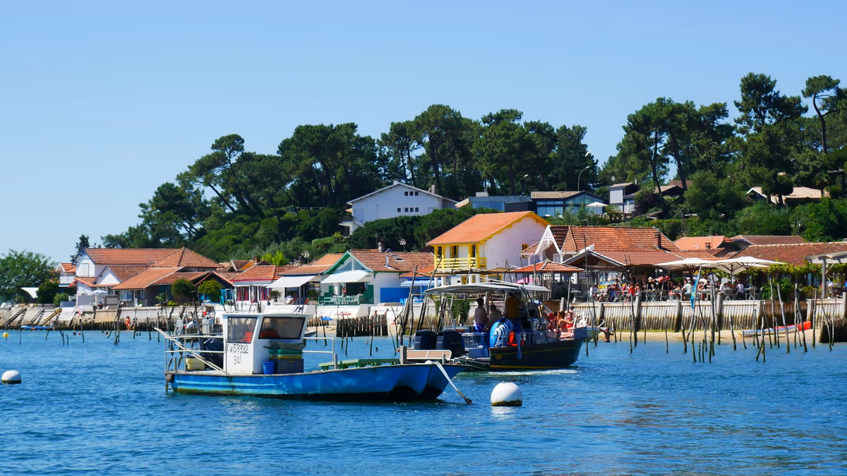Maisons aux toits de tuiles alignées au ras de l'eau dans un village du Bassin d'Arcachon, pinède en arrière-plan, bateaux de travail amarrés devant