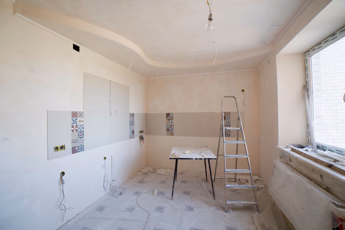 Cuisine en rénovation inachevée : faïence posée par bandes, murs nus, gaines électriques en attente et escabeau sur le sol protégé