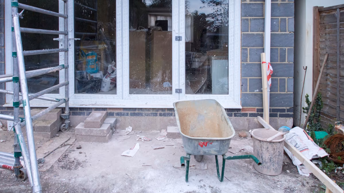 Extension de maison en travaux à l'arrêt : échafaudage roulant contre une baie vitrée neuve, brouette et seau de gâchage abandonnés sur une dalle brute