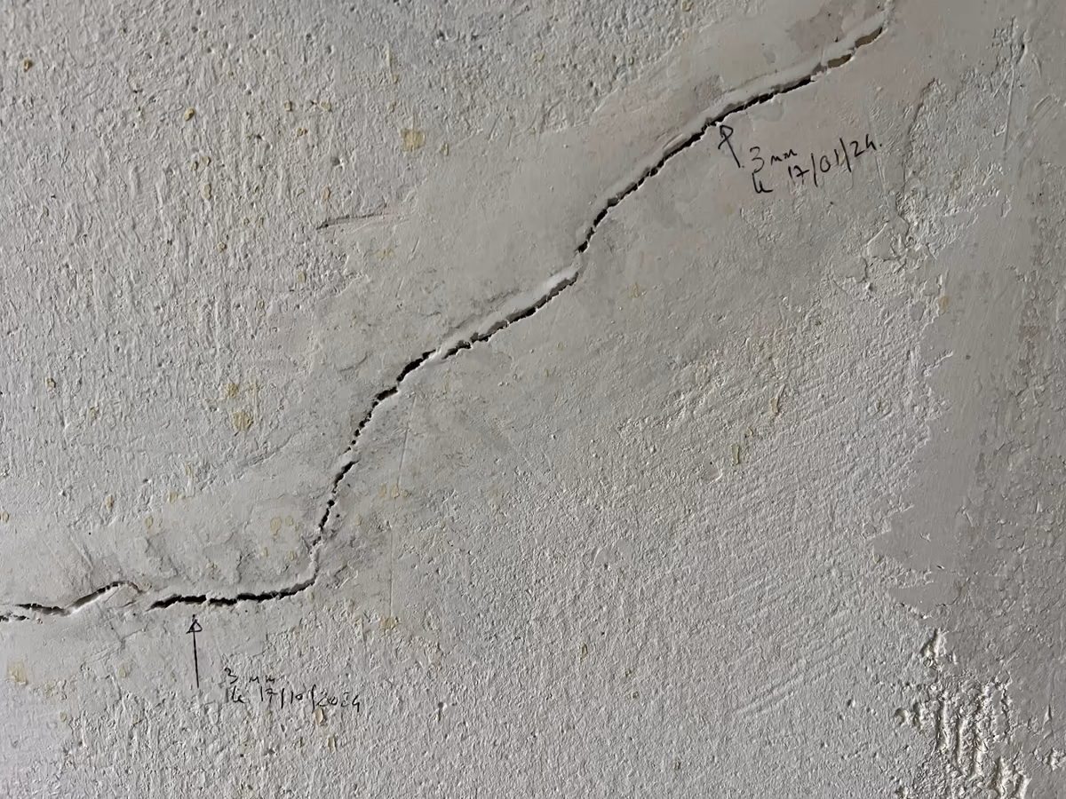 Fissure oblique dans un enduit, ses extrémités repérées au crayon avec l'ouverture mesurée et la date de chaque relevé