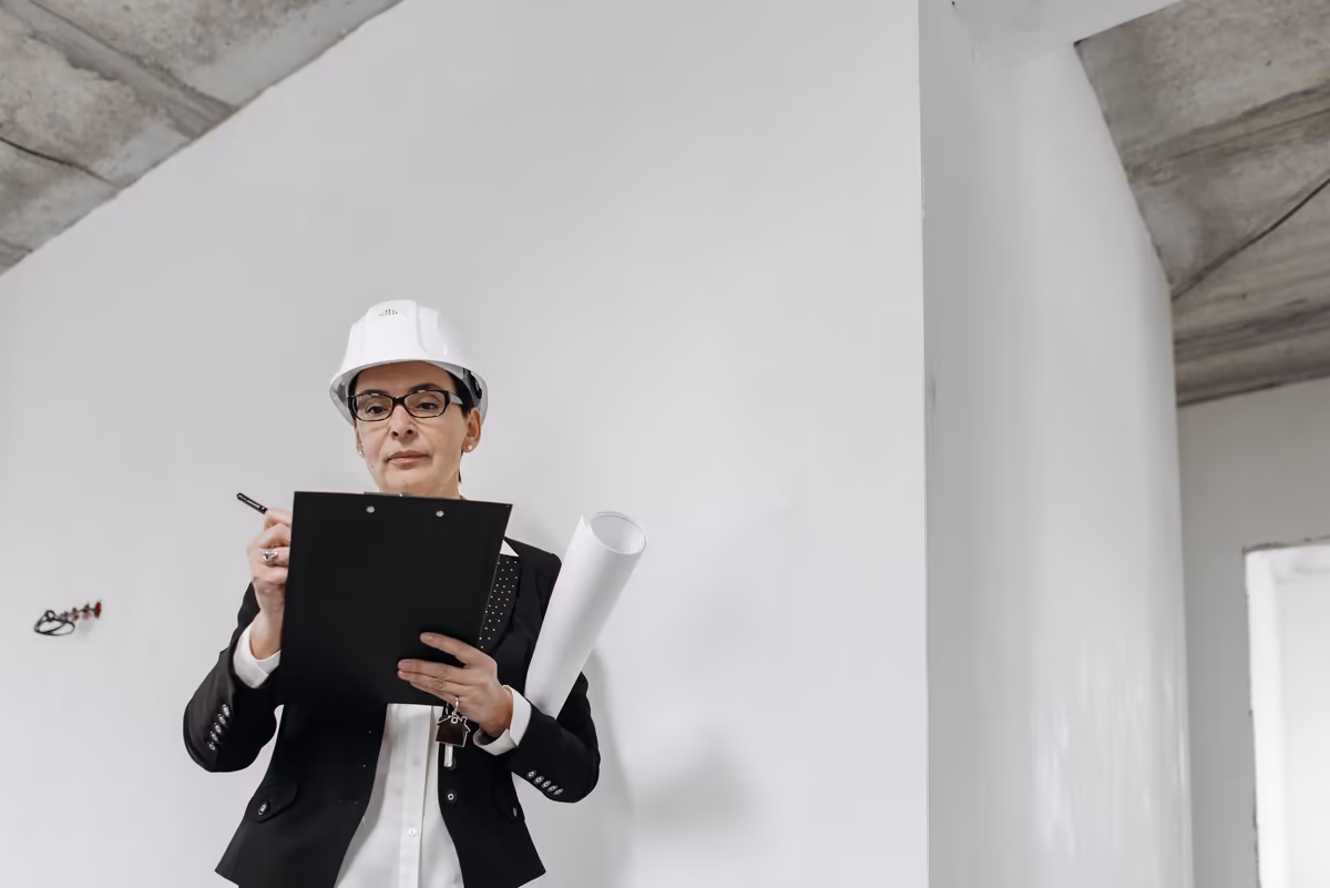 Expert en casque de chantier rédigeant son constat sur un porte-bloc, plan roulé sous le bras, dans un logement en cours de visite