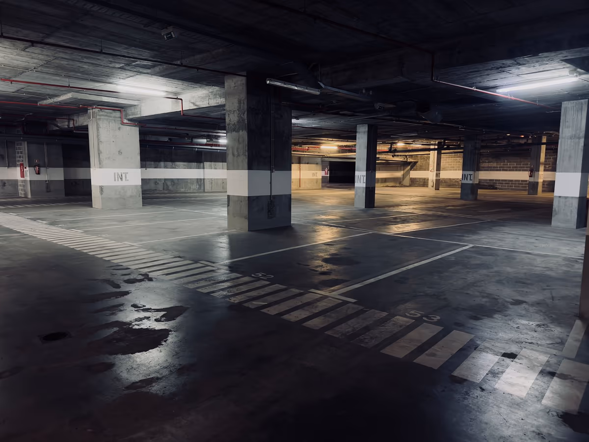 Parking en sous-sol aux poteaux en béton brut, flaques au sol et réseaux suspendus sous la dalle, faible éclairage