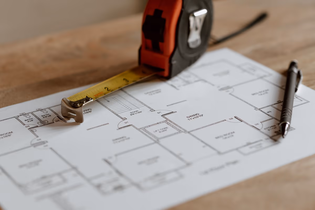 Plan de maison déplié sur une table de travail, mètre ruban déroulé posé en travers des pièces et stylo prêt à annoter une cote