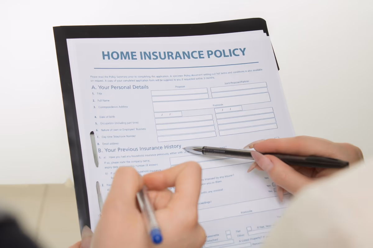 Deux mains renseignent au stylo un formulaire d'assurance habitation posé sur un porte-documents, rubriques coordonnées et antécédents visibles