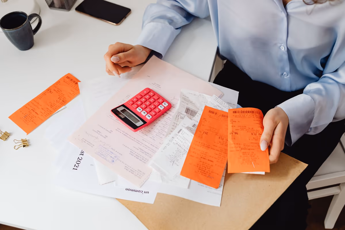 Personne assise à une table qui trie des factures et des tickets étalés autour d'une calculatrice pour établir un inventaire chiffré poste par poste