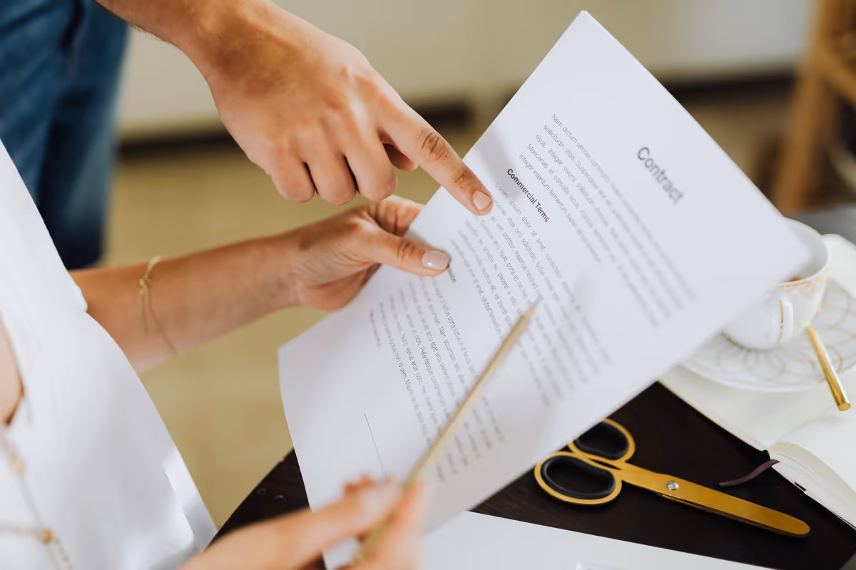 Deux personnes relisent un contrat imprimé tenu à la main, un doigt posé sur le paragraphe des conditions, stylo et carnet sur la table à côté