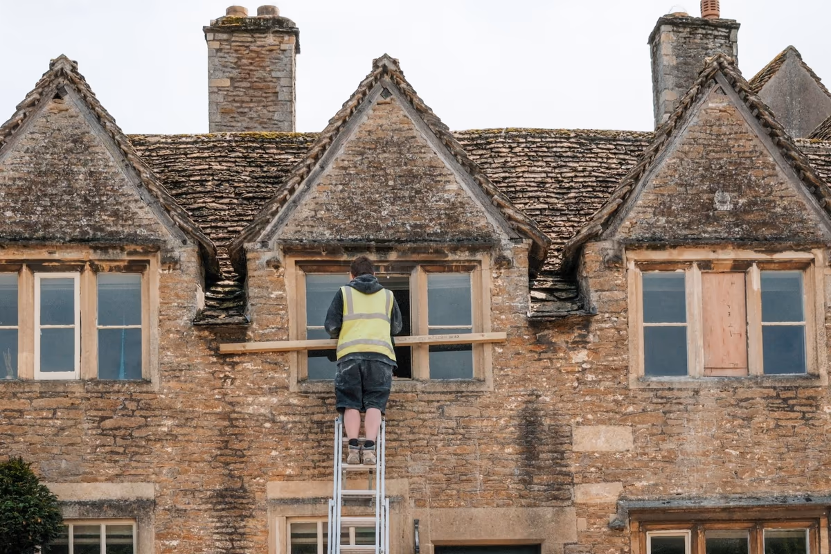 Façade en pierre d'une maison ancienne en cours de travaux, ouvrier de dos sur une échelle devant une fenêtre déposée et étayée par une pièce de bois, lucarnes et couverture en pierre au-dessus