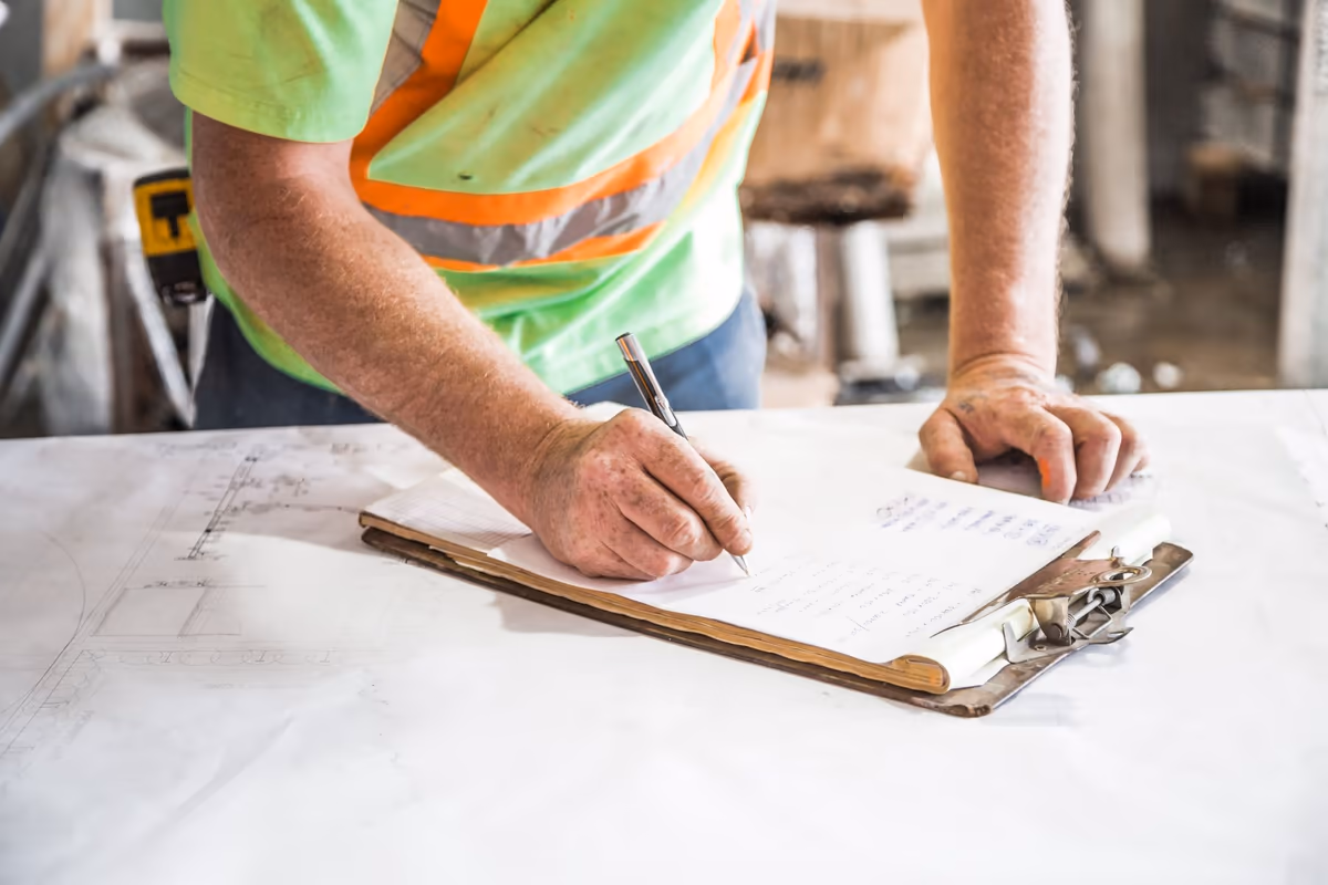 Sur un chantier, un intervenant sans visage dans le cadre écrit ses relevés au stylo sur un porte-bloc, posé sur des plans dépliés qui couvrent la table