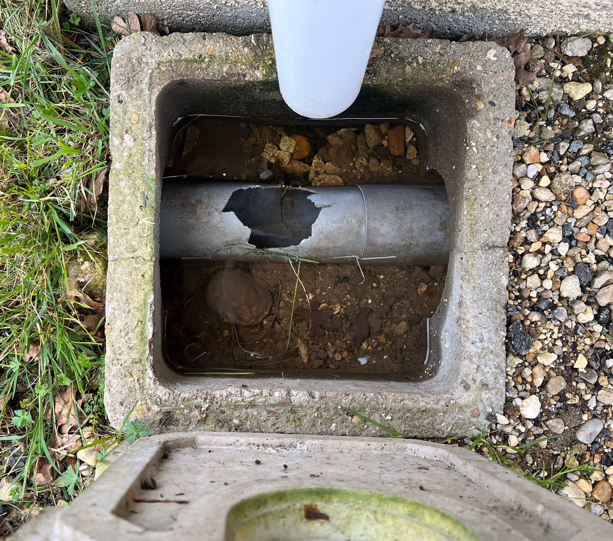 Regard d'eaux pluviales en béton ouvert sous une descente, rempli d'eau stagnante, traversé par un tuyau PVC percé, avec un fond de terre et de graviers