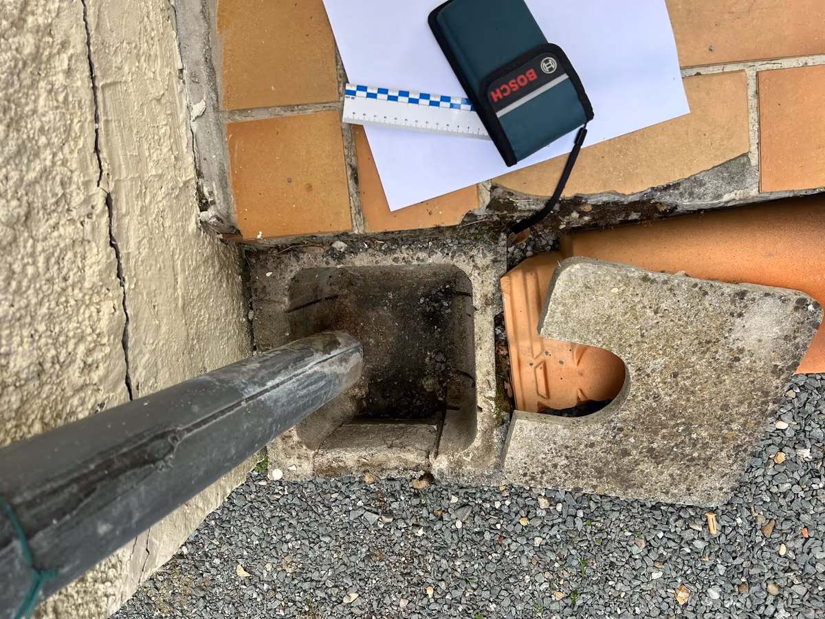Regard d'eaux pluviales ouvert au pied d'une façade enduite fissurée, sous une descente, avec télémètre laser et réglet posés sur la terrasse pour le relevé de l'expert
