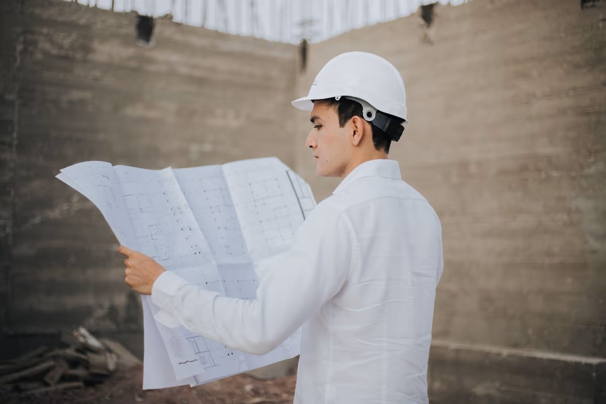 Technicien casqué lisant un jeu de plans déplié à hauteur de regard sur un chantier, devant un mur de béton brut décoffré