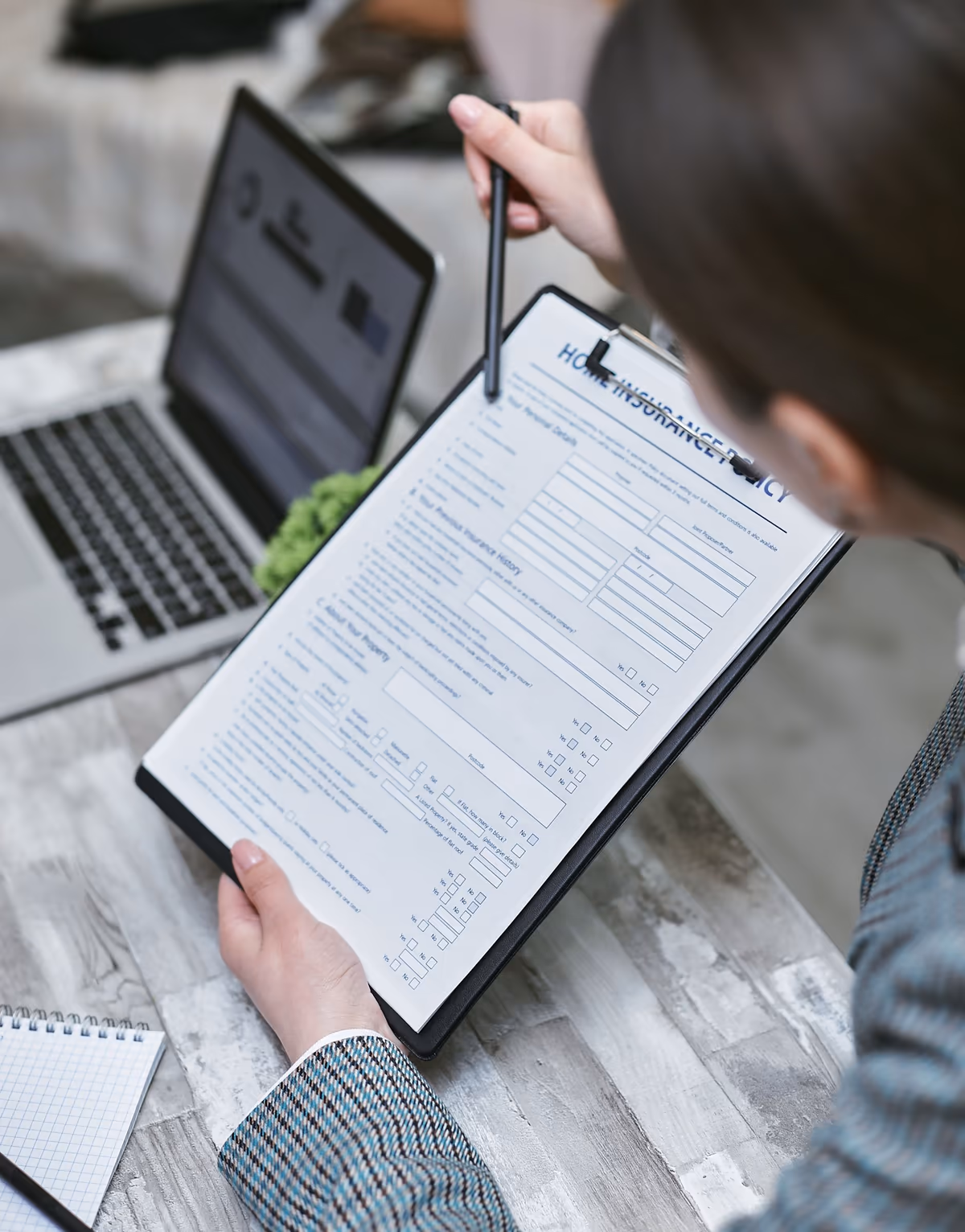 Personne relisant stylo en main un contrat d'assurance habitation fixé sur un porte-bloc, bloc-notes et ordinateur portable posés à côté sur la table