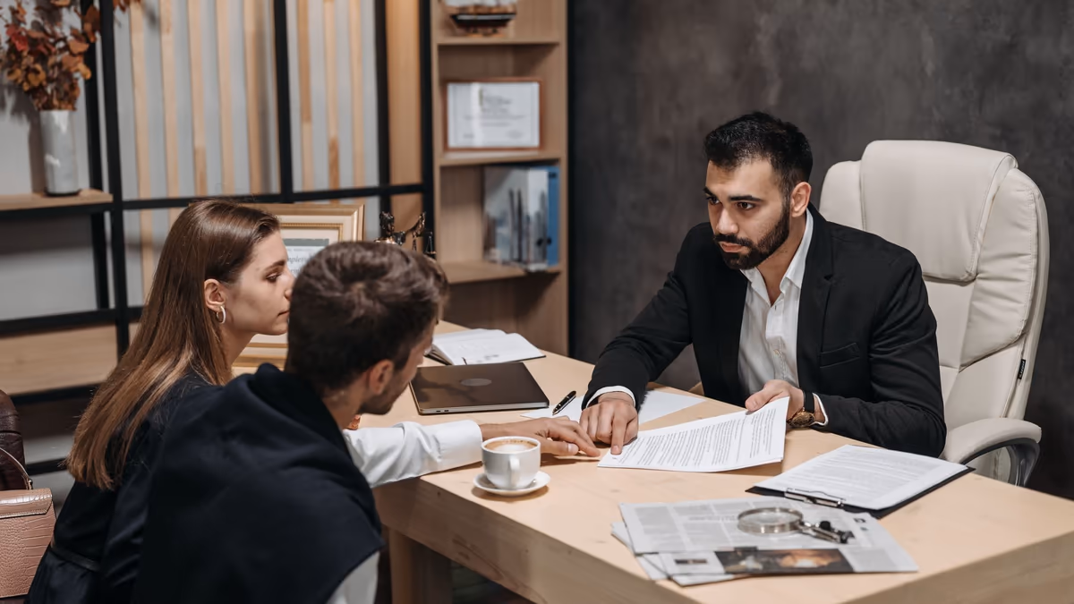 Avocat relisant un document avec un couple de propriétaires à son bureau, pièces du dossier et loupe posées sur la table, au moment de décider entre voie amiable et expertise judiciaire