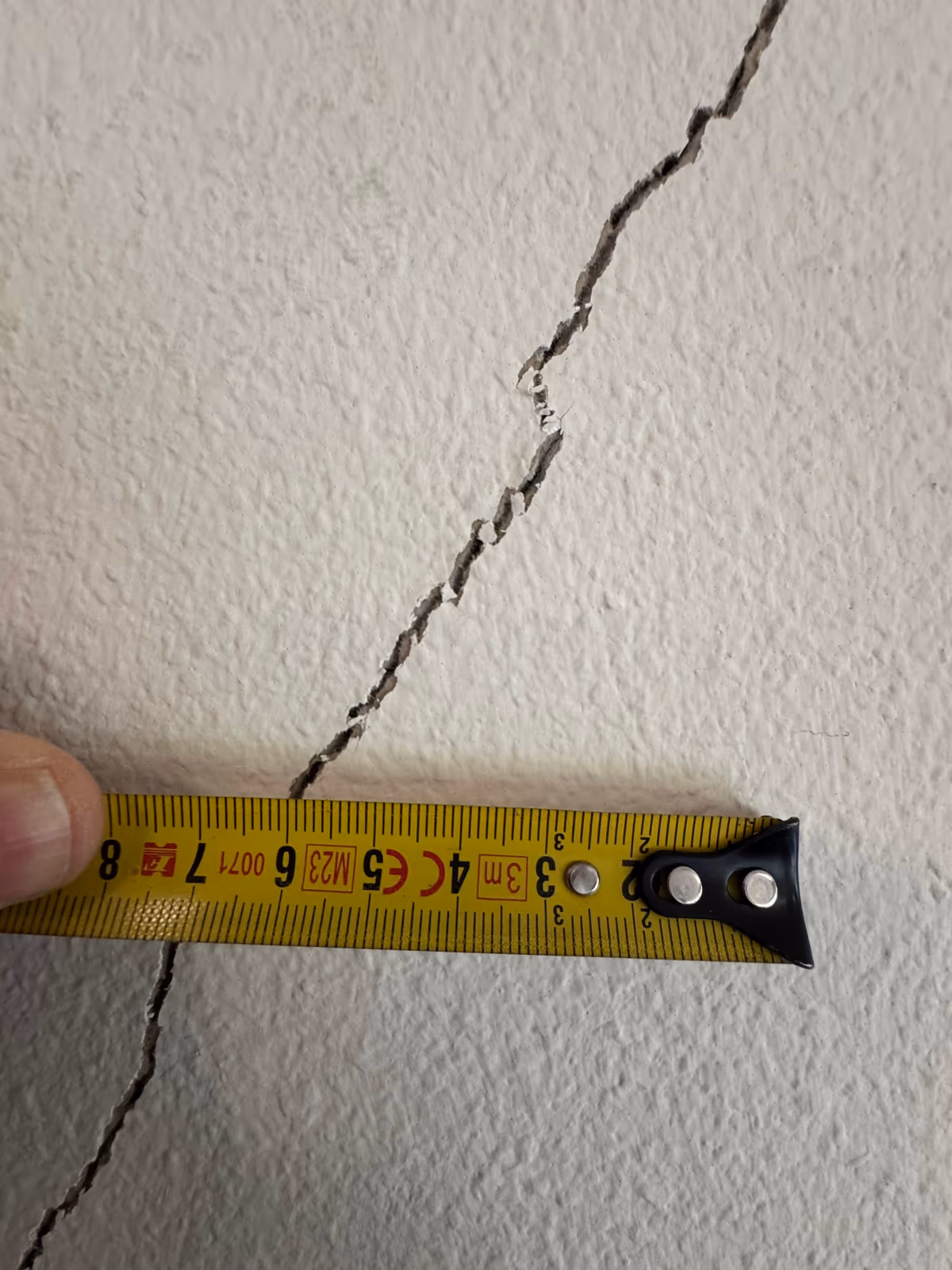 Mètre ruban tenu en travers d'une fissure oblique aux lèvres ébréchées sur un mur intérieur crépi blanc