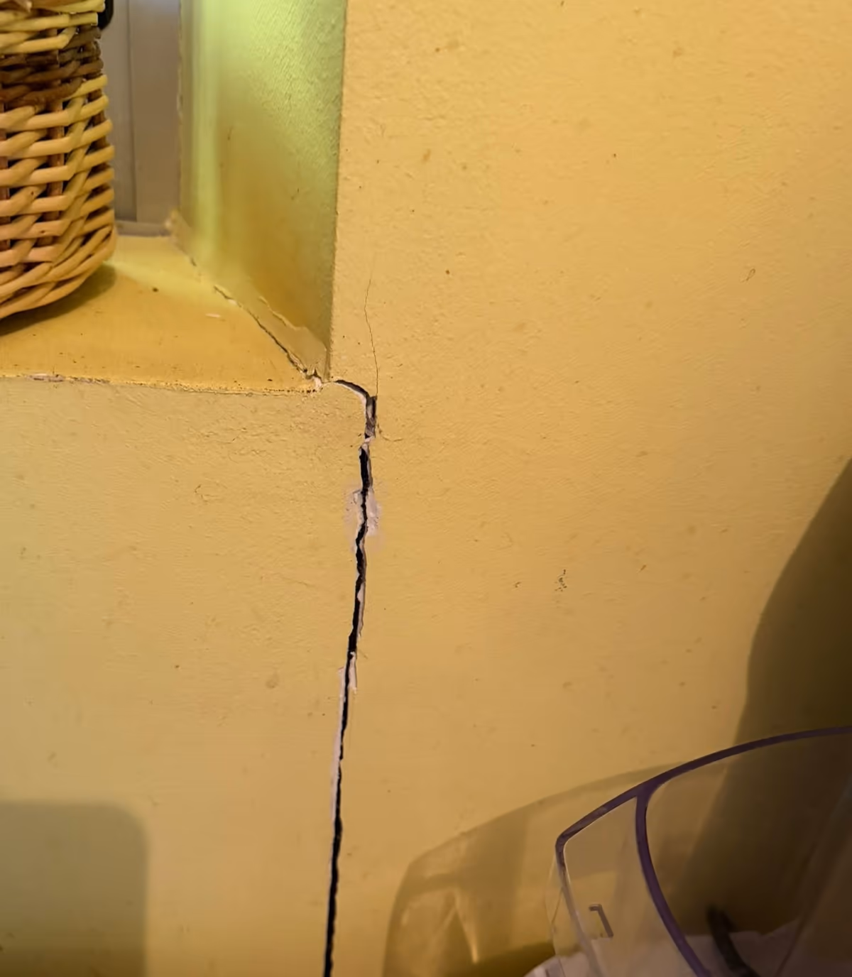 Fissure verticale ouverte partant de l'angle d'un ébrasement de fenêtre et descendant sur un mur intérieur peint en jaune