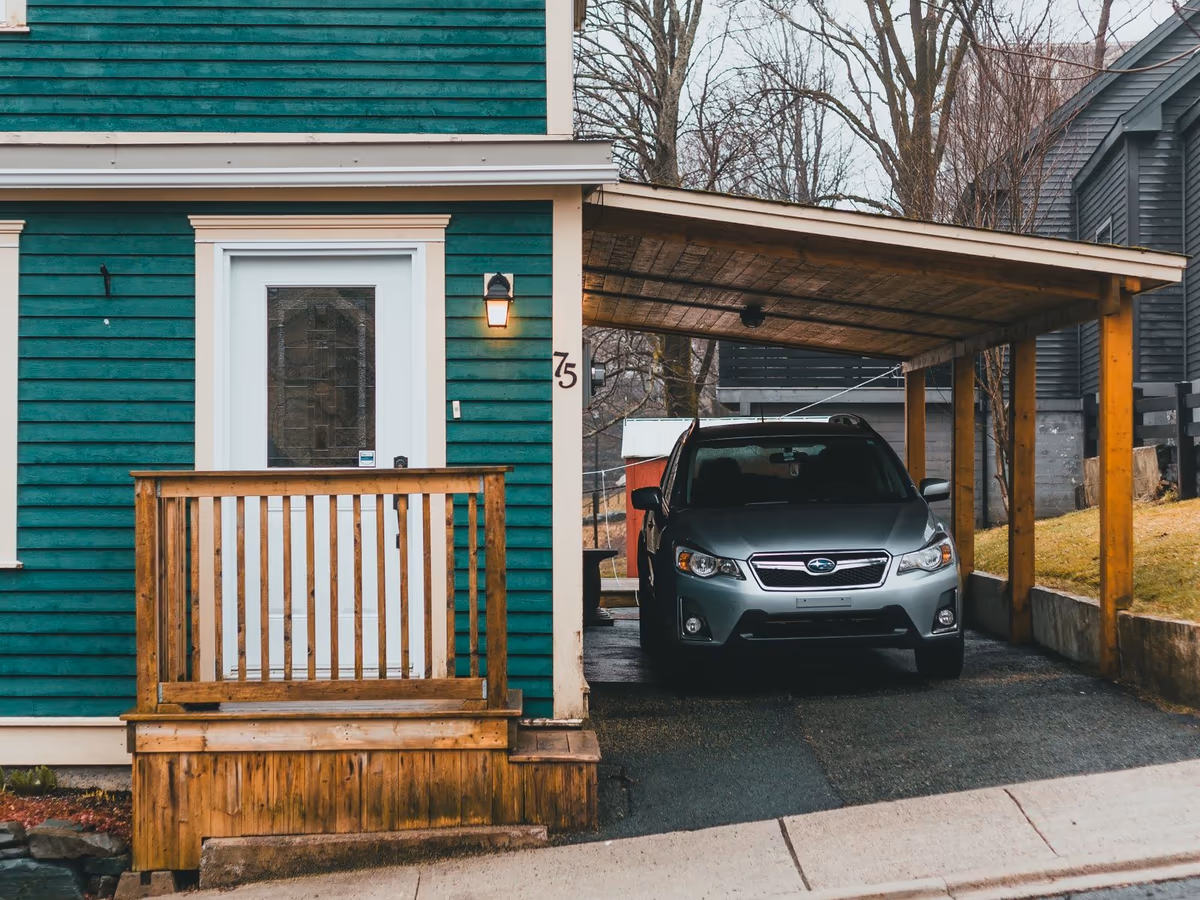 Carport accolé au pignon d'une maison individuelle en bardage bois : toiture plate portée par des poteaux posés au sol, aucun mur de remplissage, voiture stationnée dessous
