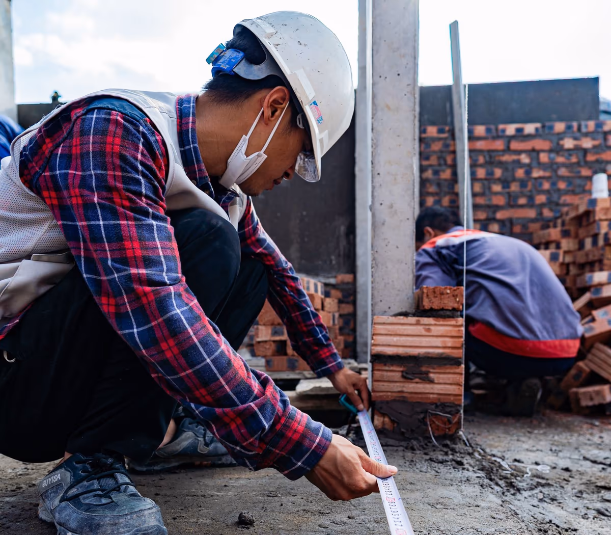 Homme casqué accroupi relevant une cote au ruban le long d'une dalle, au pied d'un mur de briques en cours de montage sur un chantier de maçonnerie