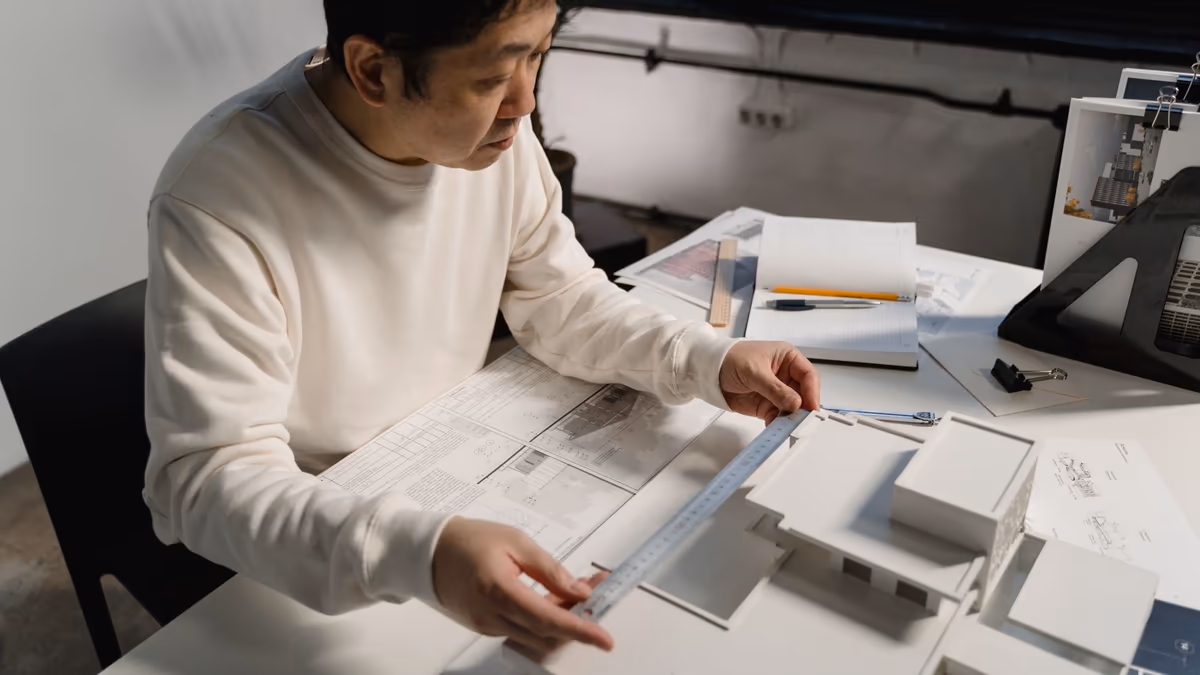 Homme mesurant au réglet la maquette blanche d'un projet de maison posée sur des plans techniques dépliés, sur une table de bureau