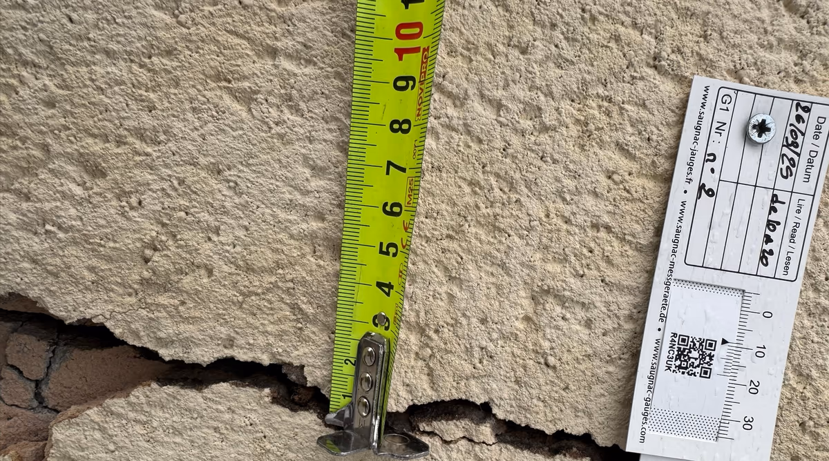 Jauge témoin vissée de part et d'autre d'une fissure ouverte dans un enduit de façade, date de pose inscrite, mètre ruban posé dans la fissure