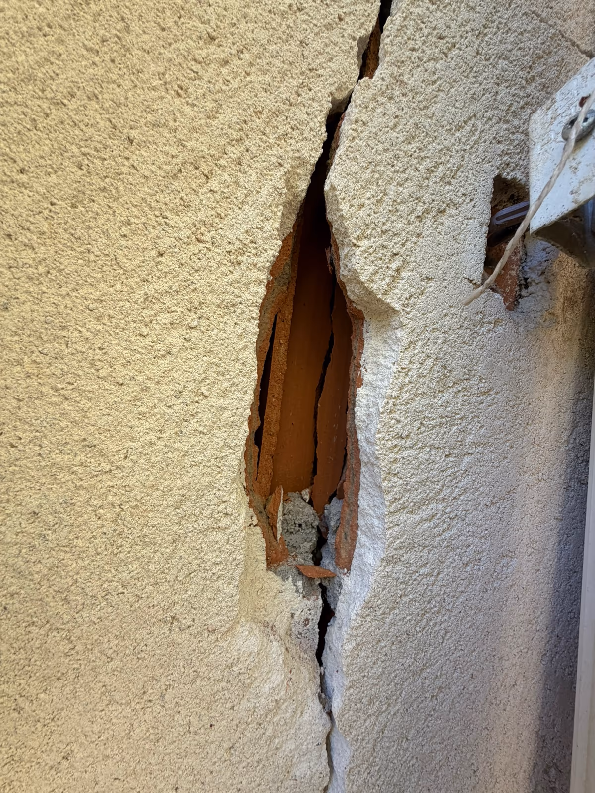 Enduit de façade éclaté le long d'une fissure verticale largement ouverte, laissant voir des briques creuses fendues dans l'épaisseur du mur