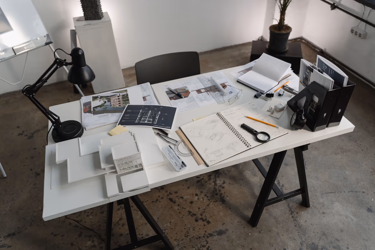Table de travail couverte d'un dossier technique : plans dépliés, planches de vues imprimées du bâtiment, carnet de croquis ouvert, loupe, réglet et classeurs à côté d'une lampe d'architecte