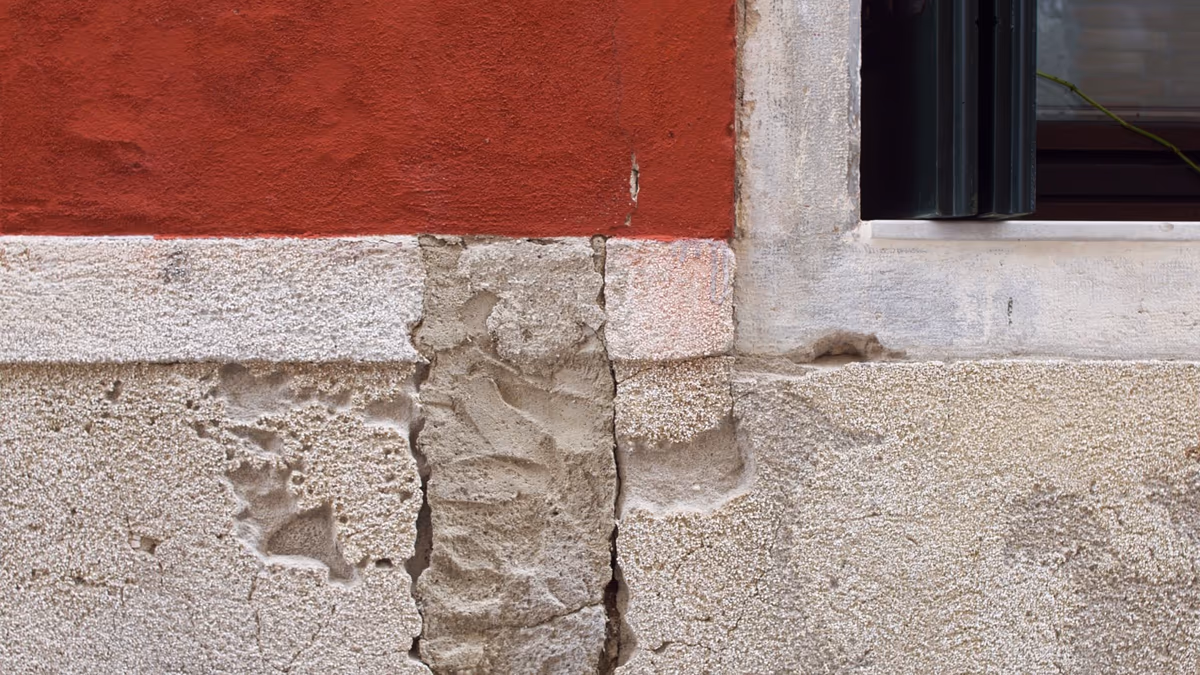 Fissure verticale ouverte dans l'enduit d'un soubassement de façade, bordée d'une bande de mortier de reprise, sous un mur peint en rouge et à côté d'un jambage de fenêtre