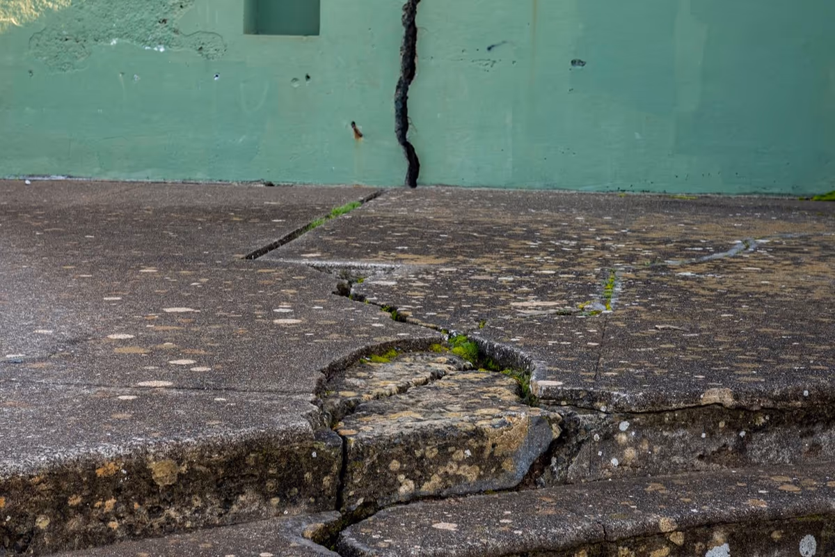 Fissure ouverte descendant un mur en béton et se prolongeant dans le dallage puis dans les marches attenantes, avec décalage des lèvres