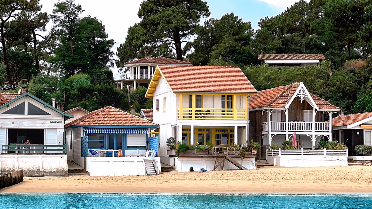 Maisons de bord de bassin en bois clair alignées face à l'eau, plage de sable au premier plan et pinède à l'arrière