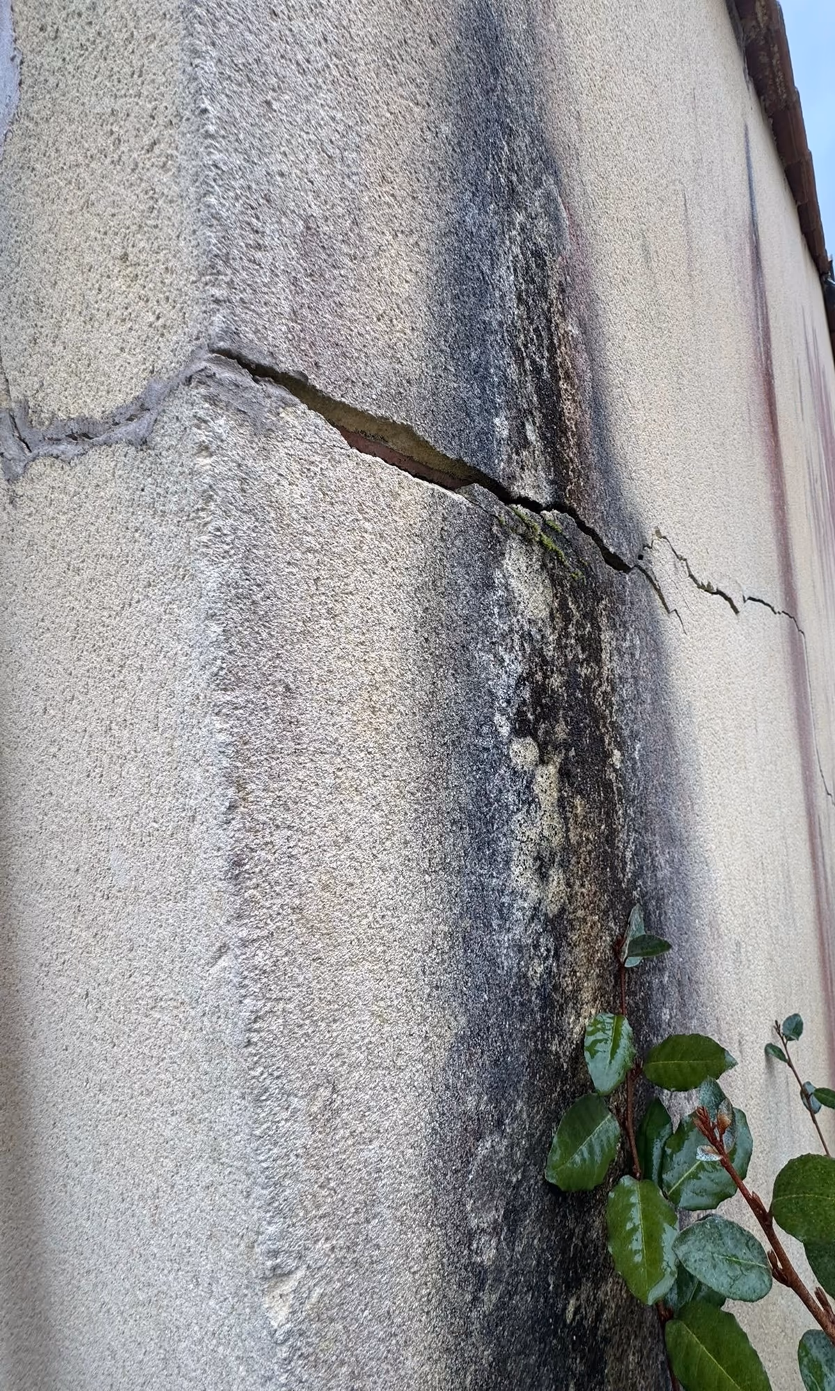 Fissure ouverte aux lèvres décalées dans l'enduit d'un angle de façade, traversée par une large traînée noirâtre verticale qui court sur l'enduit jusqu'au pied du mur