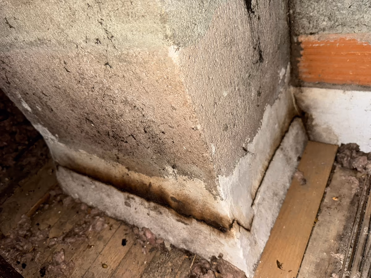 Pied d'un conduit de cheminée maçonné dans un comble : coulures brunes séchées le long de l'enduit jusqu'au plancher, isolant en vrac autour