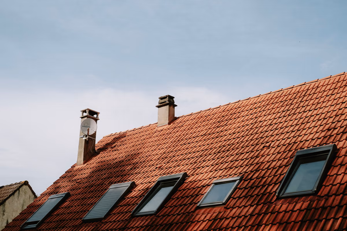 Toiture en tuiles mécaniques d'une maison individuelle sous un ciel dégagé, avec deux souches de cheminée maçonnées et quatre fenêtres de toit alignées sur le rampant