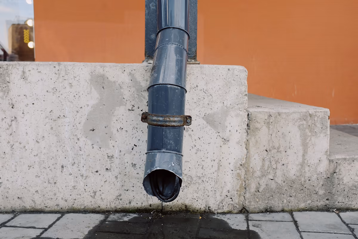 Descente d'eau pluviale en métal gris qui s'arrête juste au-dessus du sol au pied d'un mur en béton : l'eau se déverse sur le dallage contre la base du mur, mouillé et taché au point de rejet
