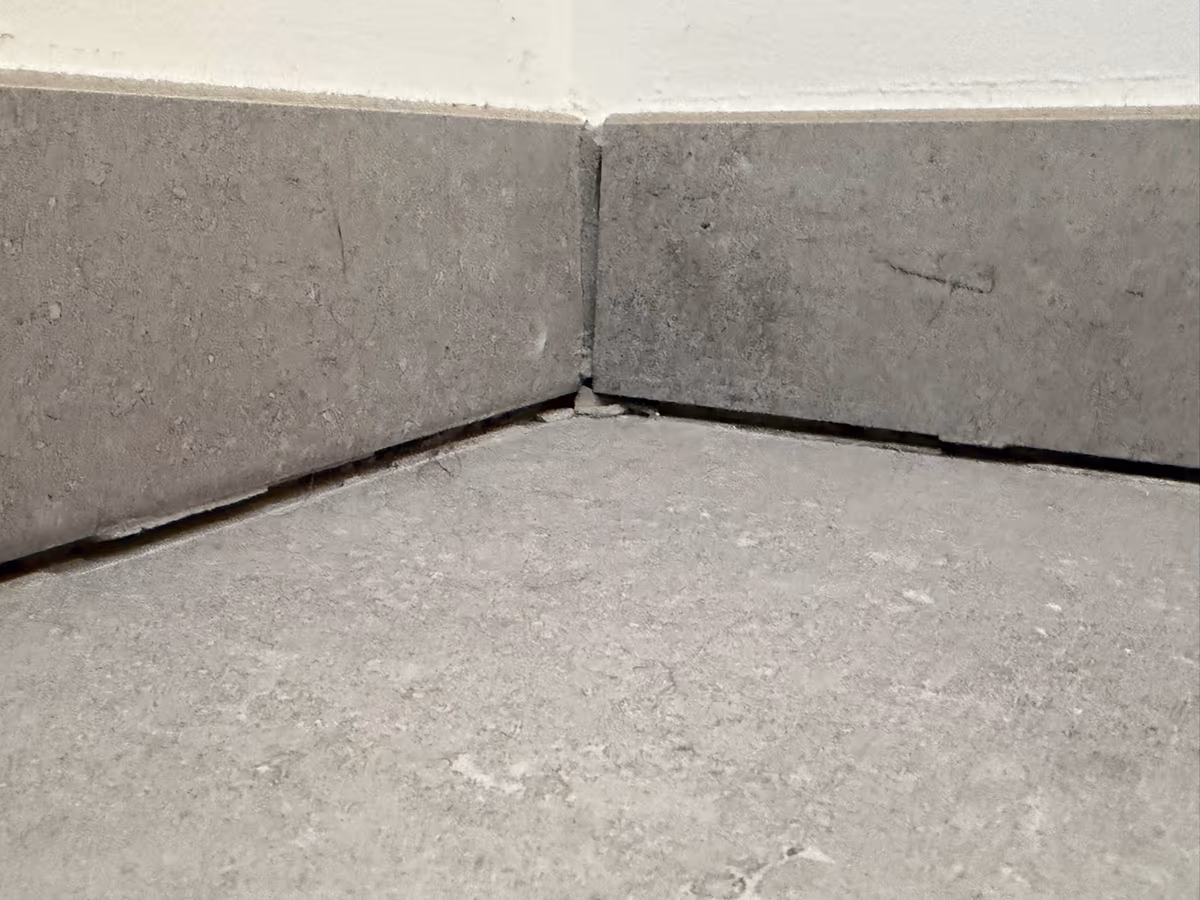 Joint ouvert entre des plinthes carrelées et le carrelage du sol, dans l’angle d’une pièce, avec une coupe d’angle mal ajustée