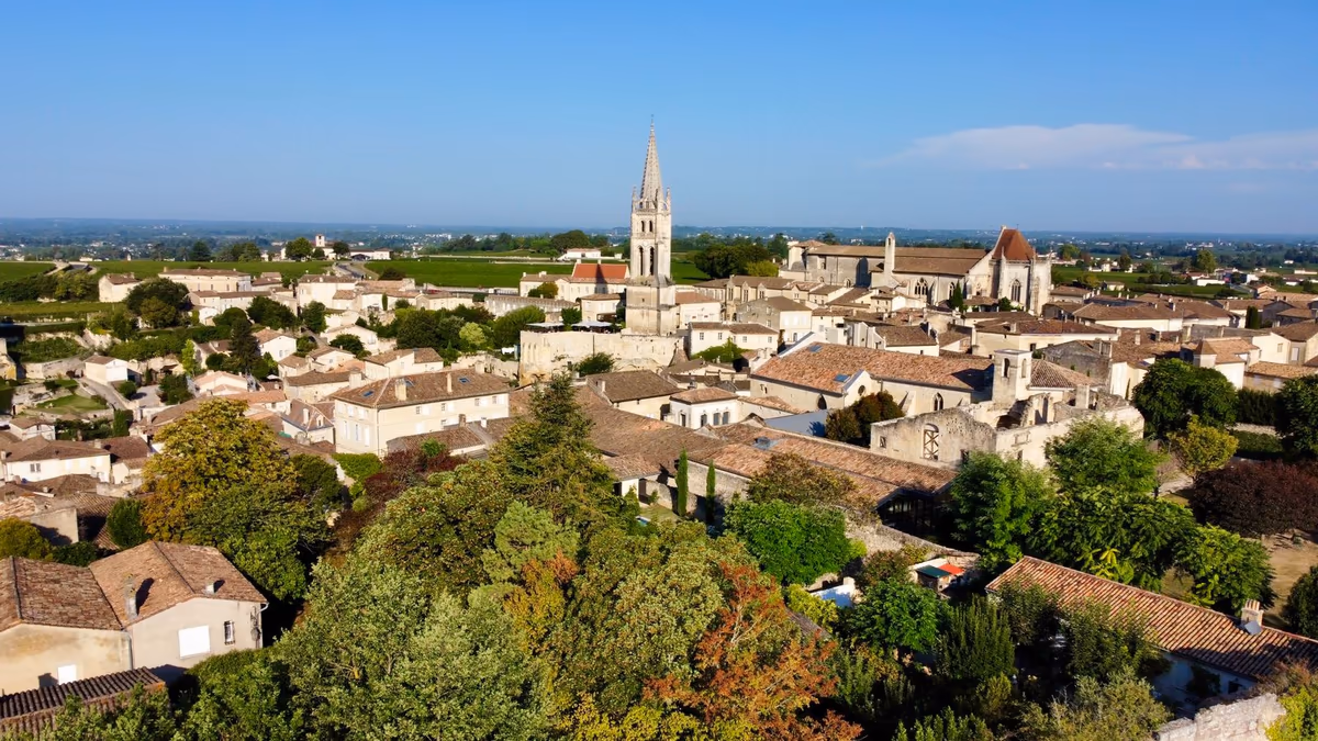 Vue en surplomb du bourg ancien de Saint-Émilion, dans le Libournais : maisons de pierre calcaire aux toitures de tuiles serrées autour du clocher, et vignes de coteau jusqu'à l'horizon