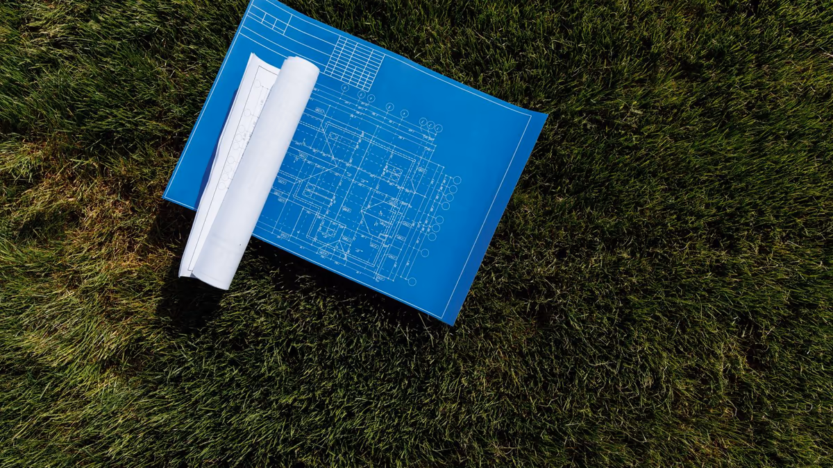 Plan d'architecte déplié et rouleau de plans posés sur l'herbe d'une parcelle, vus de dessus
