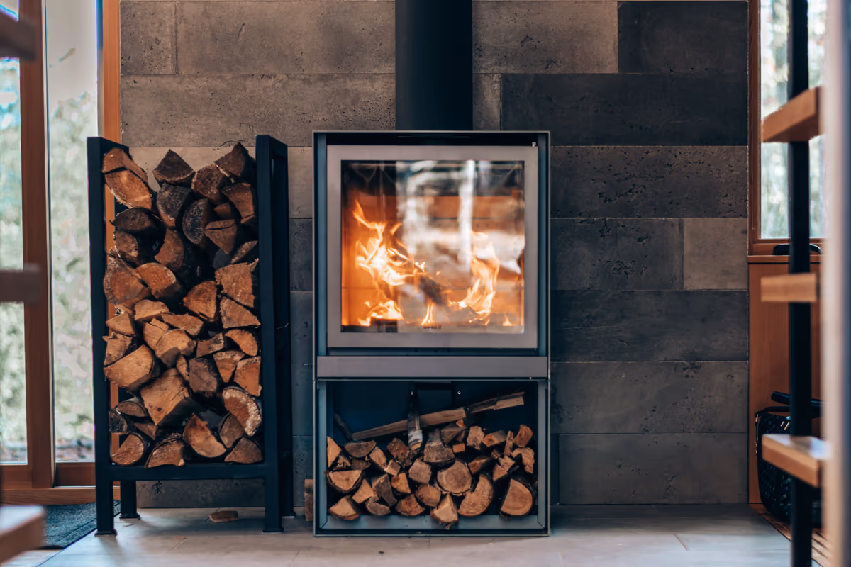 Appareil de chauffage au bois à foyer fermé et porte vitrée, installé dans le séjour d'une maison devant un parement de pierre, bûches rangées sous le foyer et sur un support métallique à côté