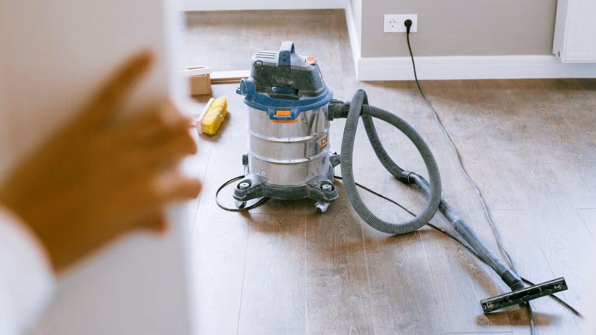 Aspirateur de chantier et balai posés au sol d’un logement rénové, sol stratifié et plinthes en place, avant remise des lieux