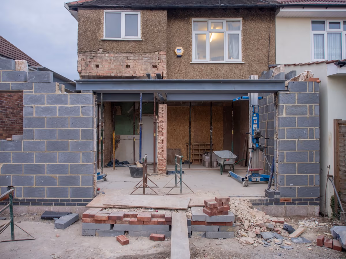 Extension de plain-pied en cours de construction à l'arrière d'un pavillon existant : murs en blocs béton montés jusqu'à l'arase, poutre métallique posée en jonction avec la façade et dalle coulée