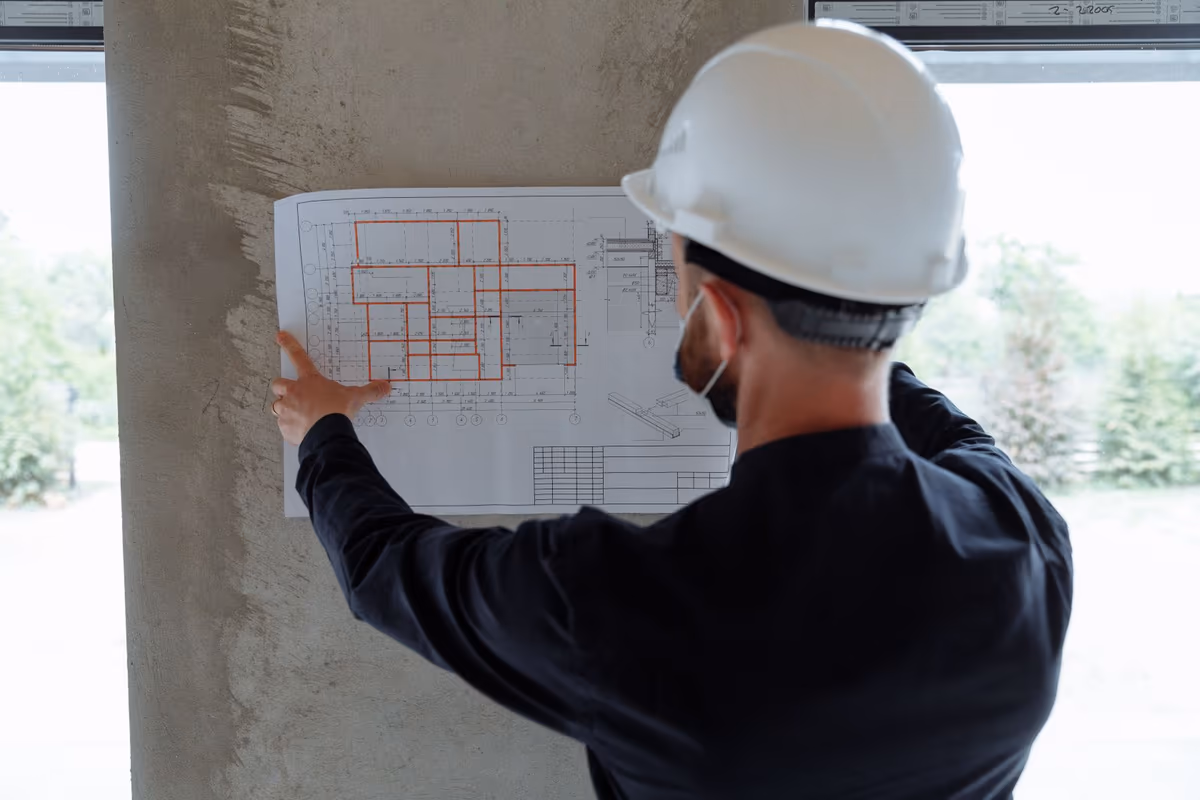 Homme casqué de dos sur un chantier, tenant un plan d'exécution déplié contre un poteau béton pour comparer le document à l'ouvrage construit