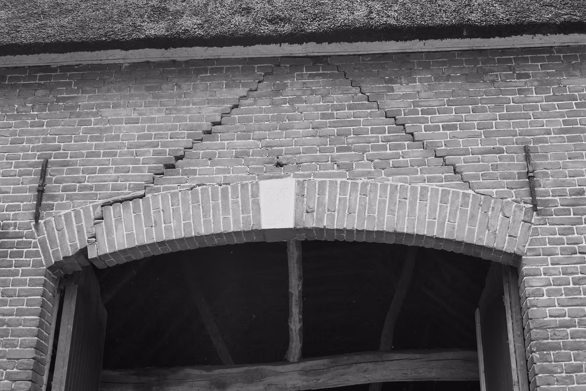 Façade en brique au-dessus d'une large porte cintrée : deux fissures en escalier suivent les joints de mortier et remontent en diagonale depuis l'arc, dessinant un triangle dans la maçonnerie