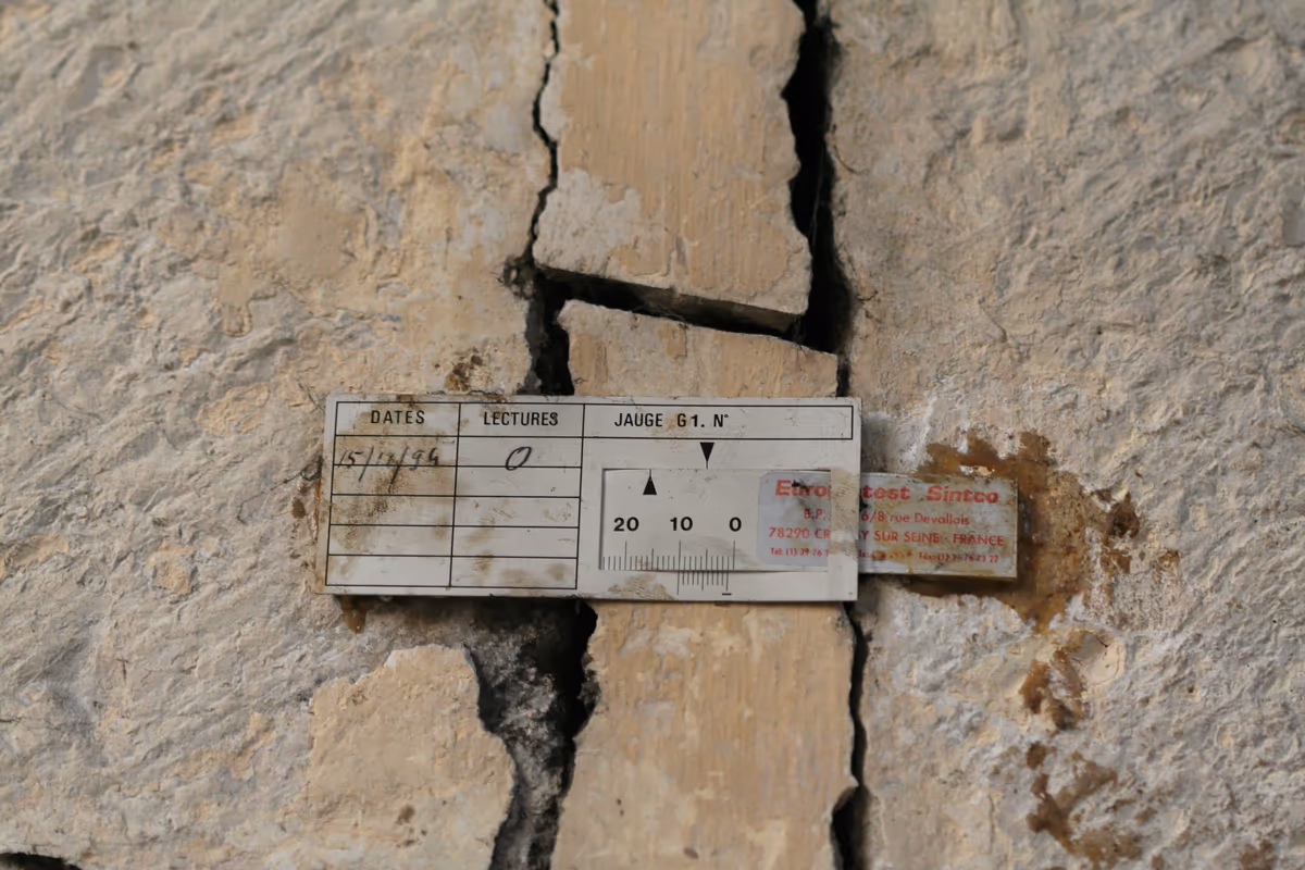 Jauge de fissure fixée à cheval sur une large fissure d'un mur en pierre, avec sa réglette graduée et une fiche à deux colonnes, dates et lectures, dont la première ligne porte le 15/12/94 et la lecture 0