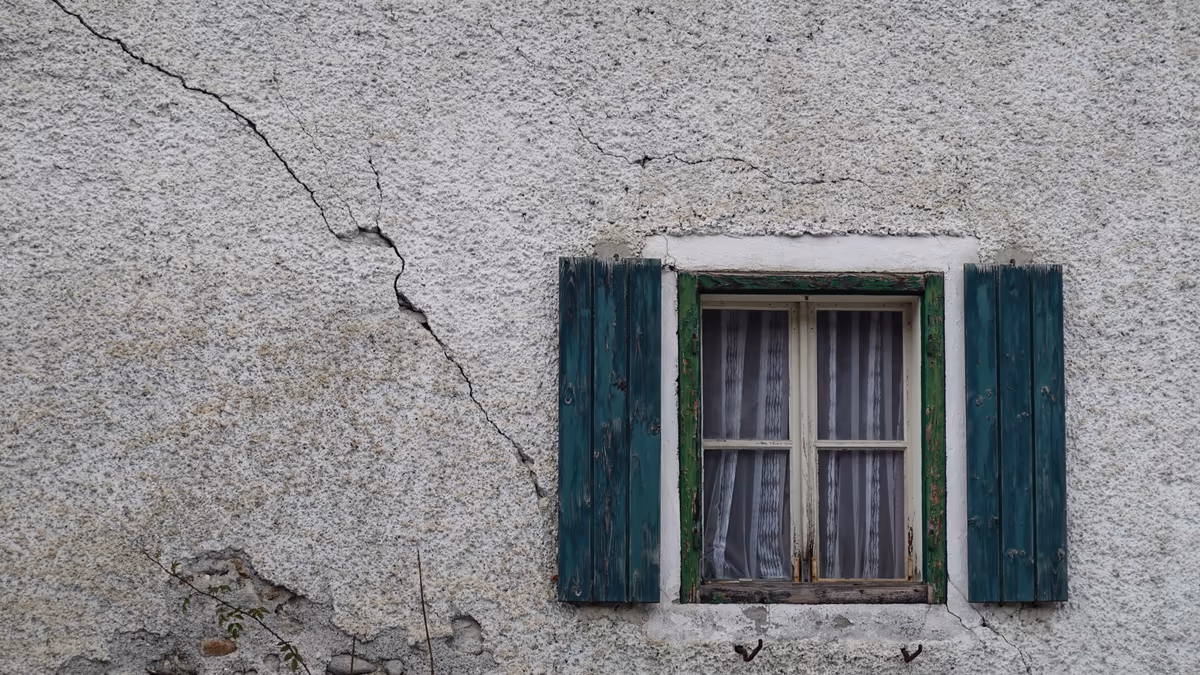 Façade enduite d'une maison ancienne traversée par une longue fissure biaise qui descend vers une fenêtre à volets verts, enduit tombé en pied de mur laissant voir la maçonnerie