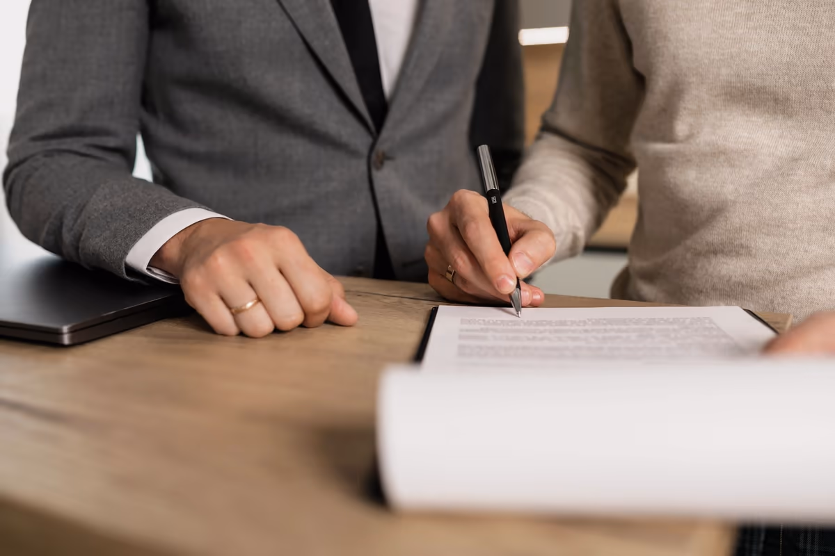 Deux personnes attablées lors d'une signature : une main tient le stylo au-dessus du document, une liasse de feuilles posée devant elles sur le plateau de bois