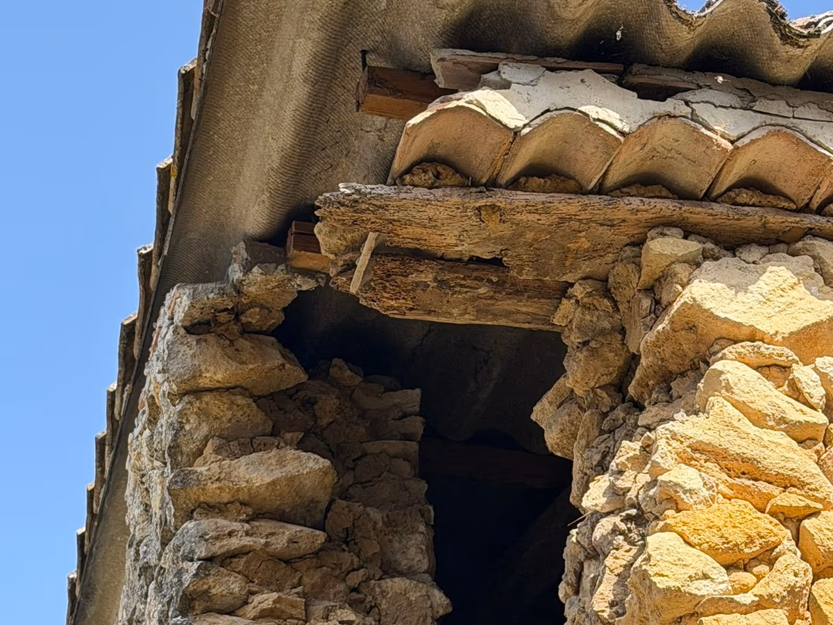 Angle d'un bâtiment ancien en pierre sous une rive de tuiles : le linteau en bois au-dessus d'une ouverture est creusé de galeries et rongé sur toute sa face visible