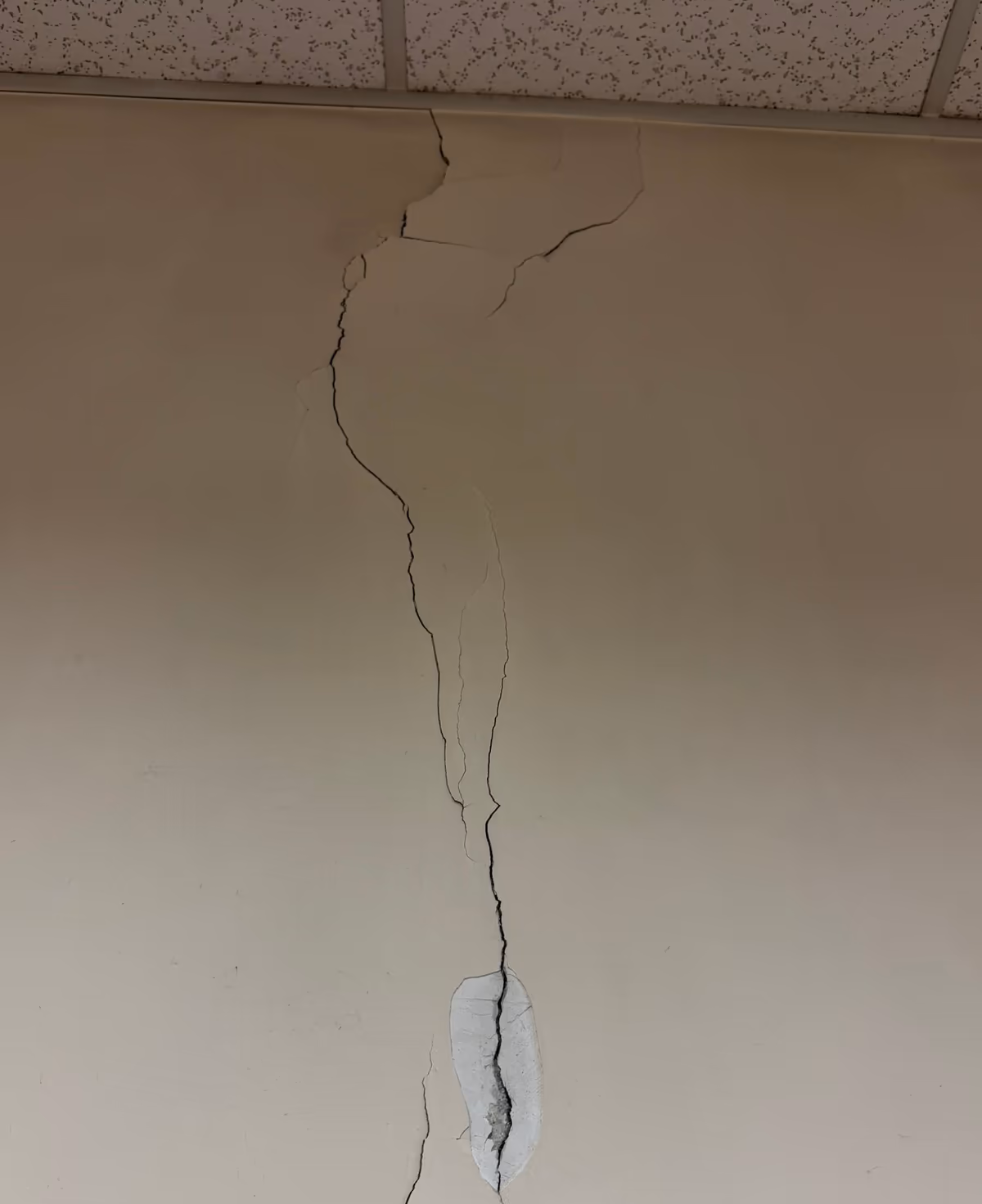 Fissure verticale sinueuse sur un mur intérieur peint, ouverte jusqu'au plafond et traversant une petite zone d'enduit reprise