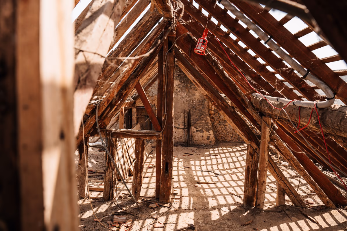 Combles non aménagés d'une maison ancienne, charpente et liteaux apparents, gravats et câbles au sol, sans plancher praticable