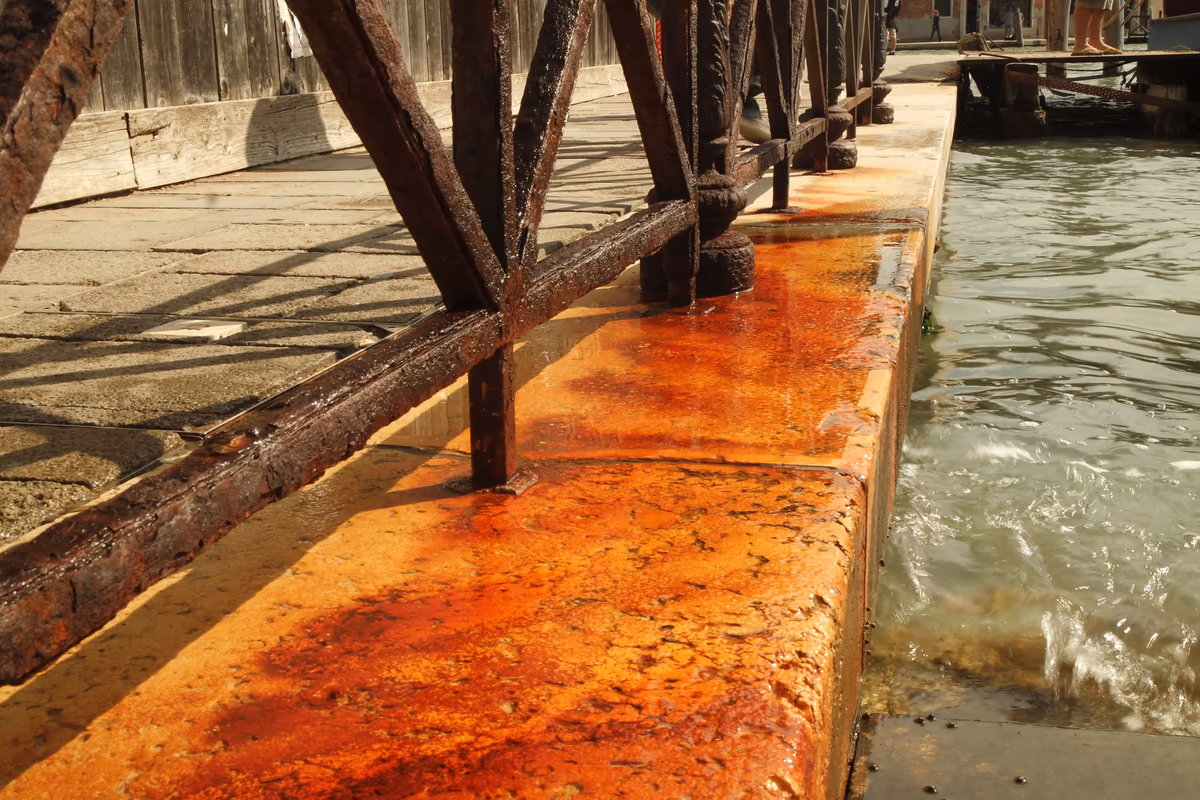 Garde-corps en fer rouillé scellé dans un bord de quai en pierre au ras de l'eau salée, coulures de rouille orangées étalées sur la pierre au pied des barreaux