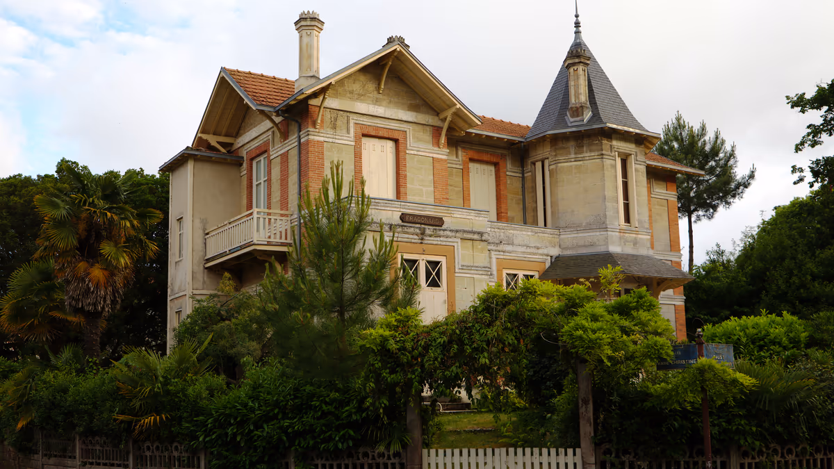 Villa Fragonard à Arcachon : façade en pierre claire et brique, balcon en bois sur consoles, tourelle d'angle coiffée d'ardoise, jardin planté de pins et de palmiers